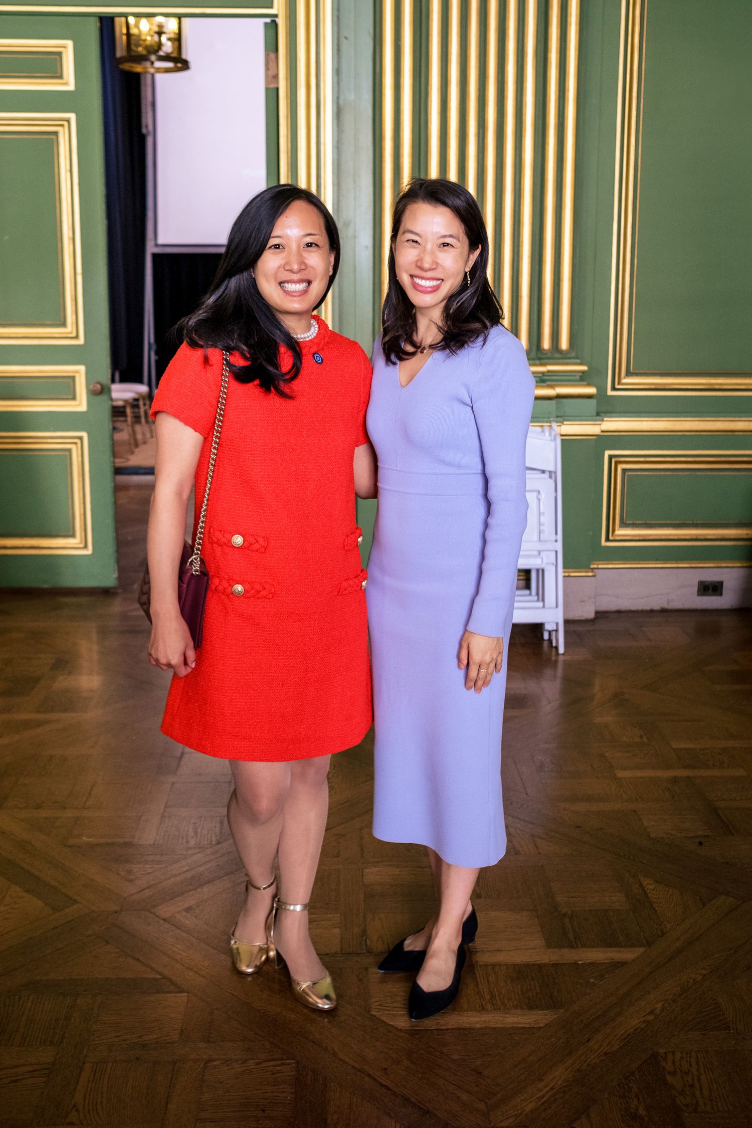 Two smiling women pose together, one in a red dress and one in a lavender midi dress, in an elegant green and gold room.
