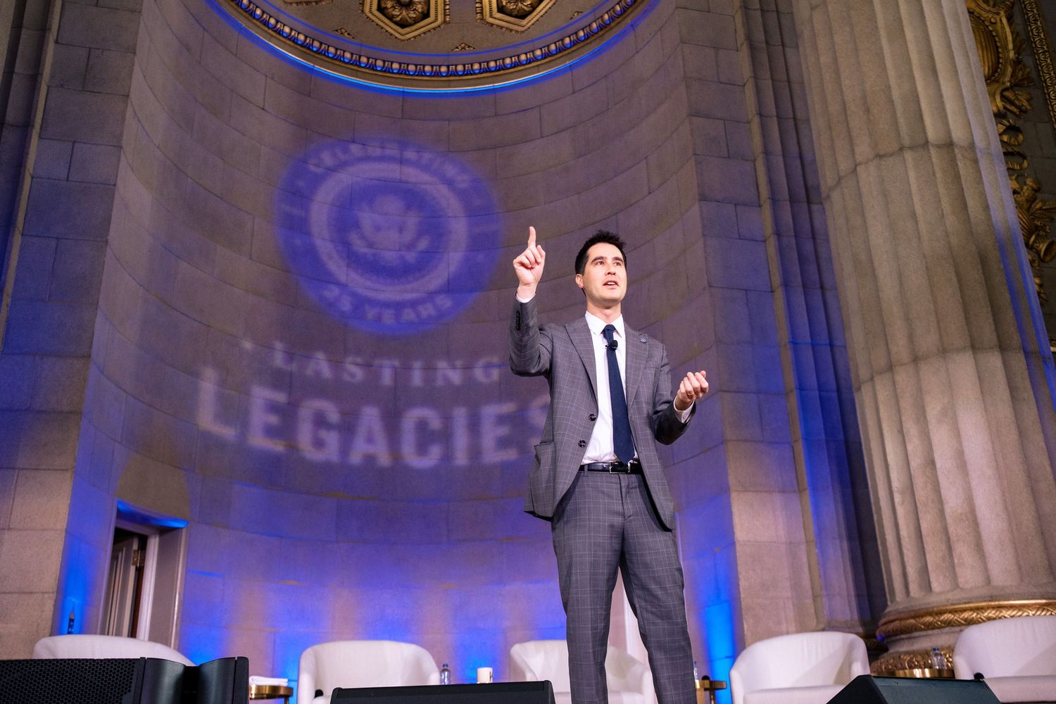Young man in gray suit speaks on stage at Lasting Legacies event beneath grand columns with blue lighting.