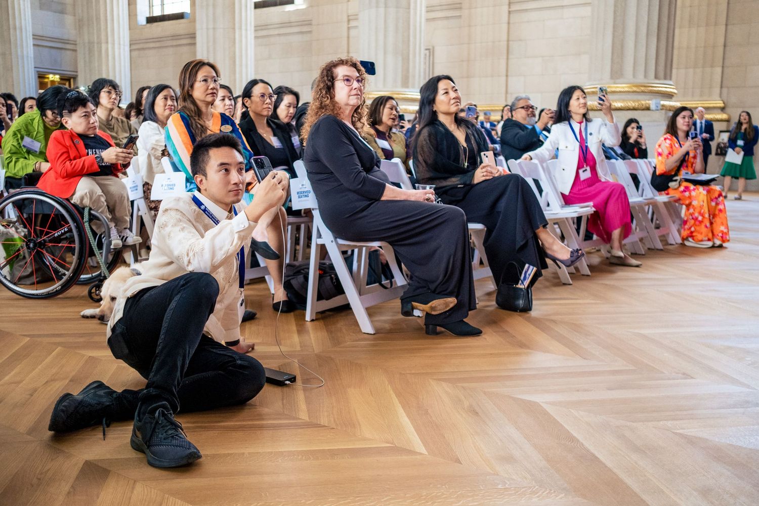 Diverse audience seated in elegant hall, including wheelchair user, watching presentation at formal multicultural event.