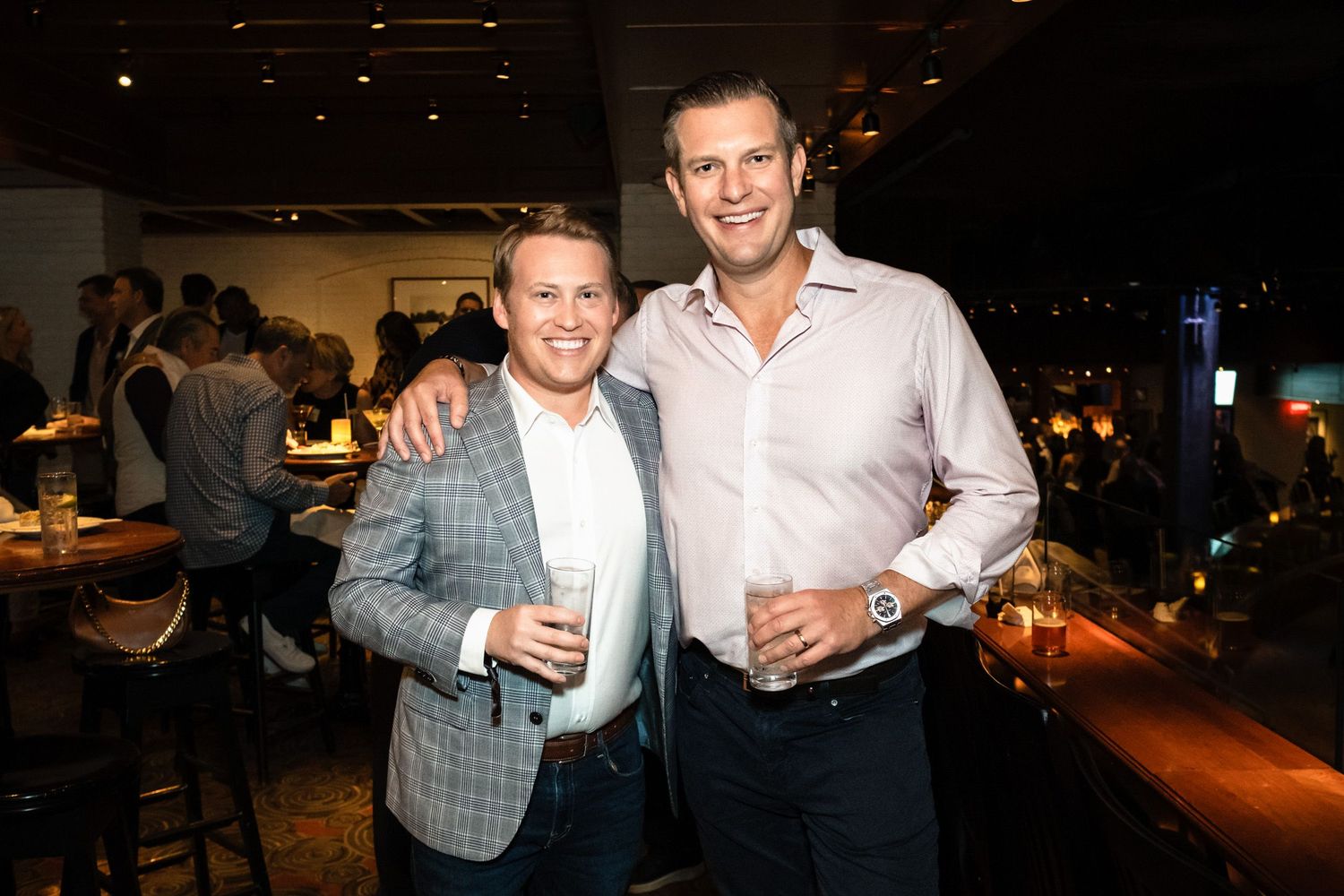 Two smiling men holding drinks pose together at a stylish bar during an evening networking event.