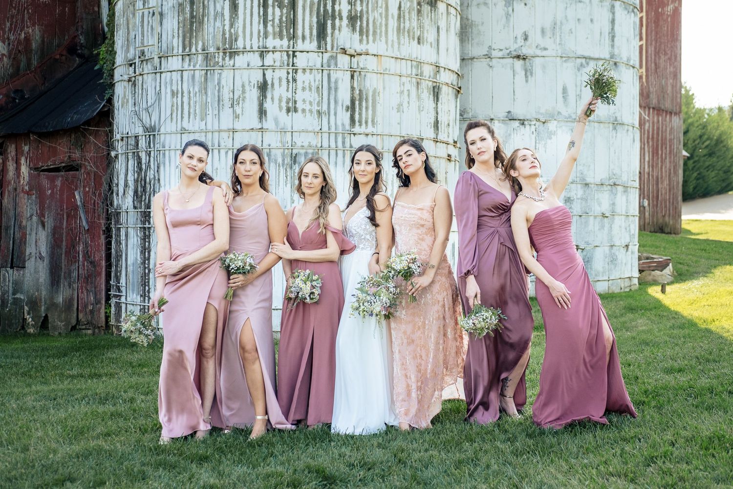 Bride poses with six bridesmaids in mismatched pink and mauve gowns beside a grain silo.