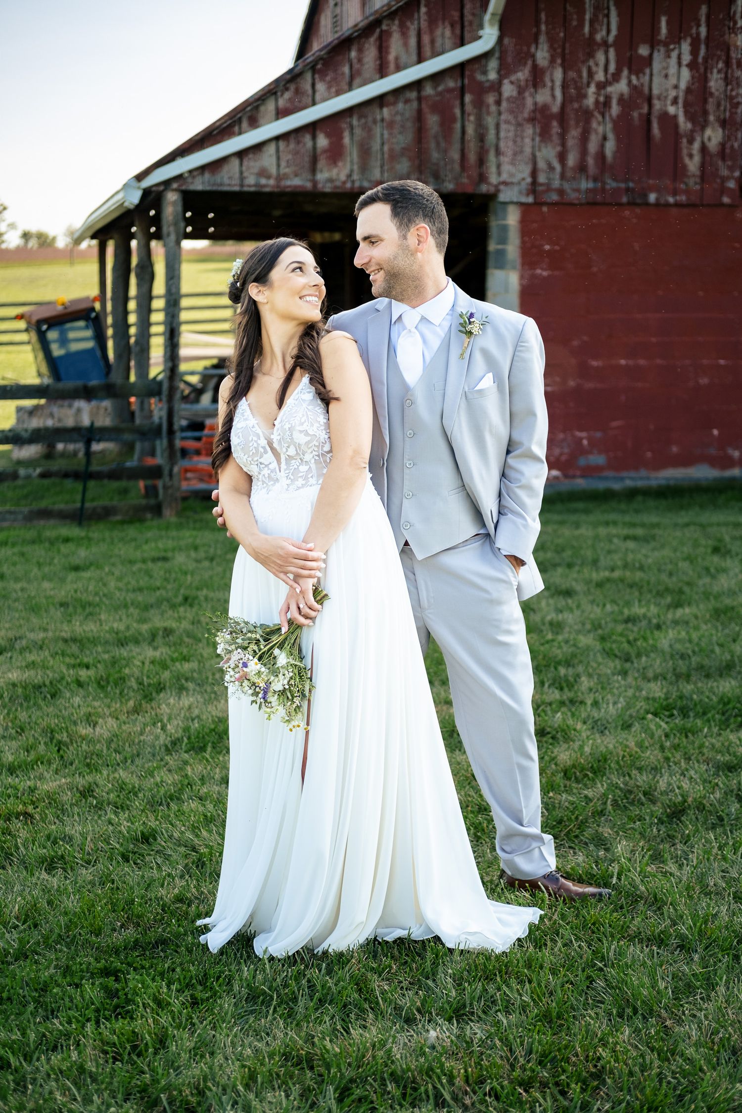 Bride and groom share a romantic moment on green lawn in front of a rustic red barn.