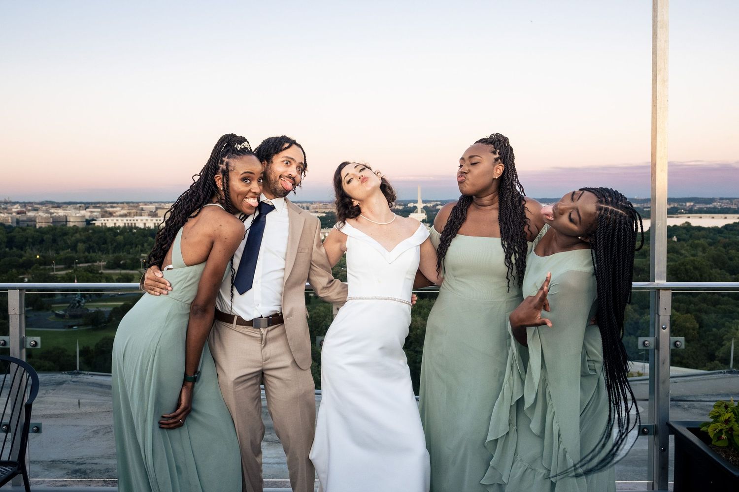 Bride poses joyfully with four bridesmaids in sage green gowns on a rooftop at sunset.