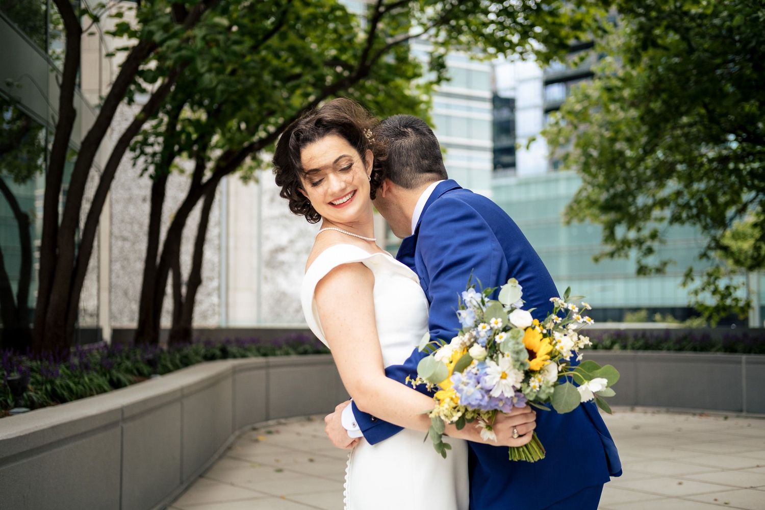 Groom in blue suit embraces bride from behind as she smiles holding a colorful wedding bouquet.