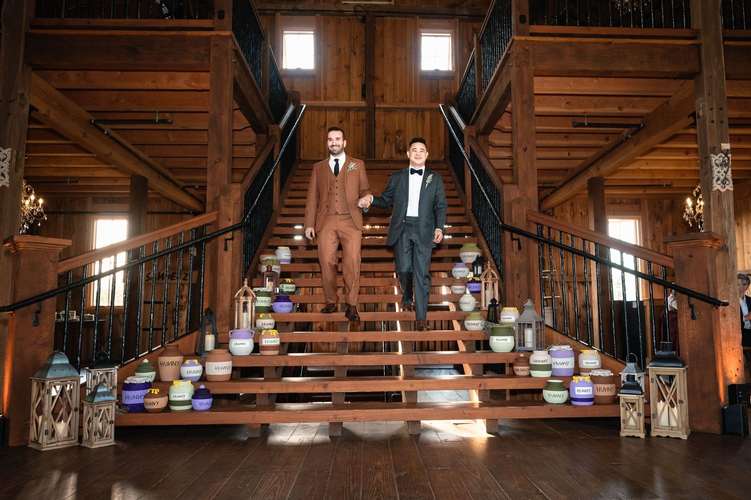 Two grooms in suits pose on decorated barn staircase at rustic wedding reception.