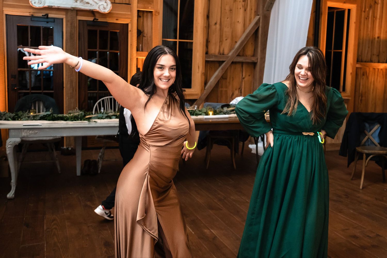 Two women in elegant gowns dance joyfully at a rustic barn wedding reception.