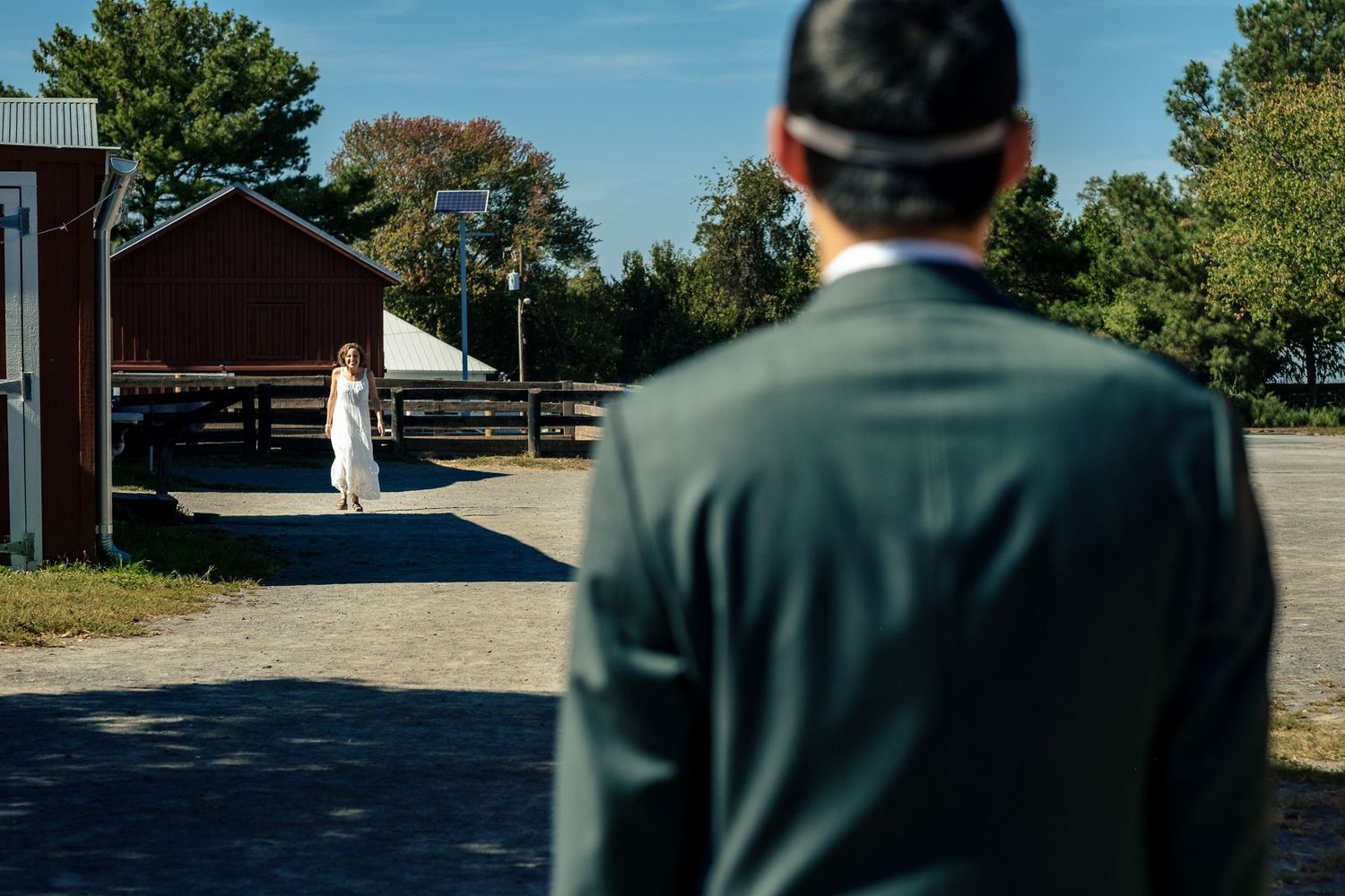 Groom in dark suit watching bride in white dress approach red barn for first look.