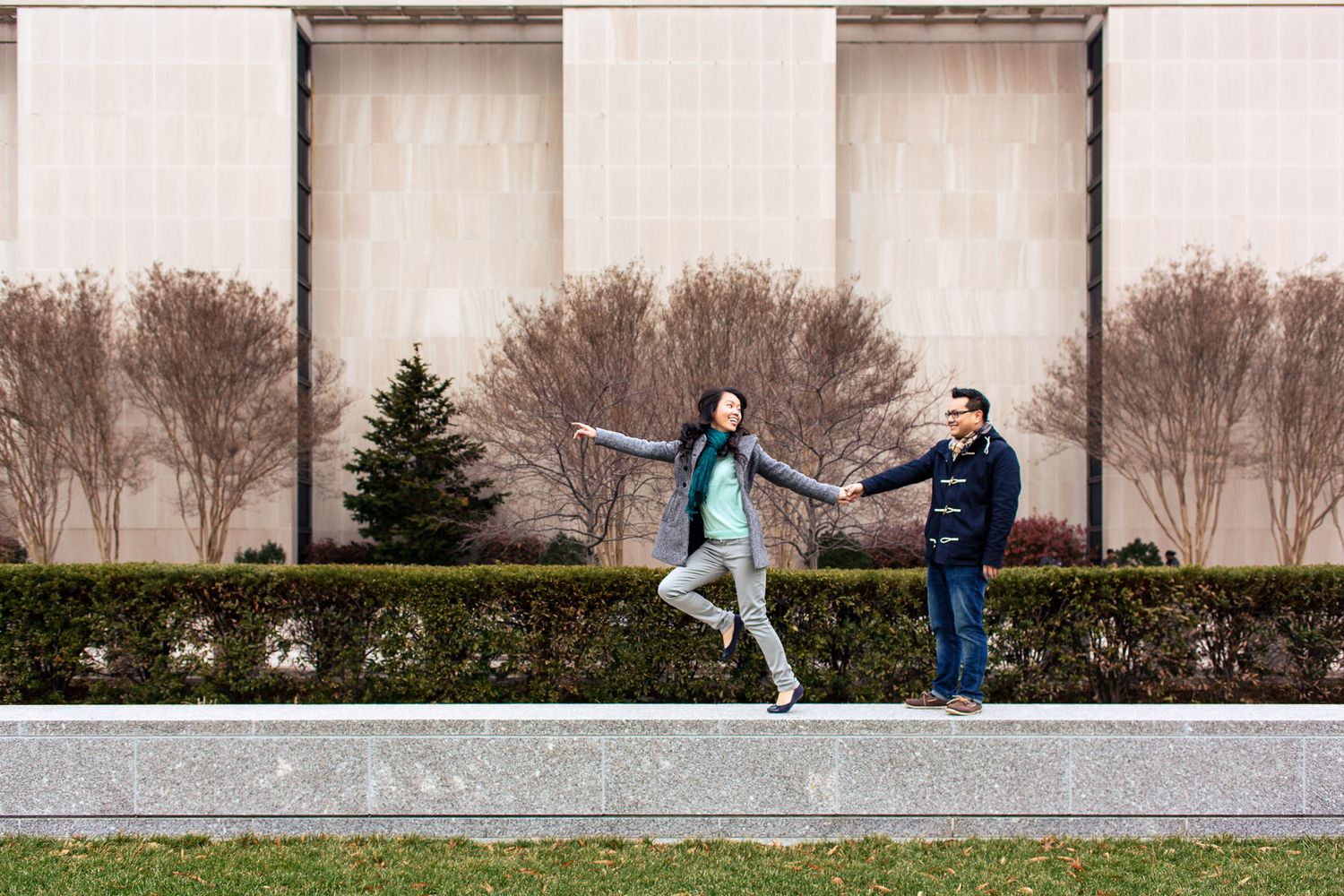 Playful couple holding hands outdoors, woman balancing on ledge near manicured hedges and large building.