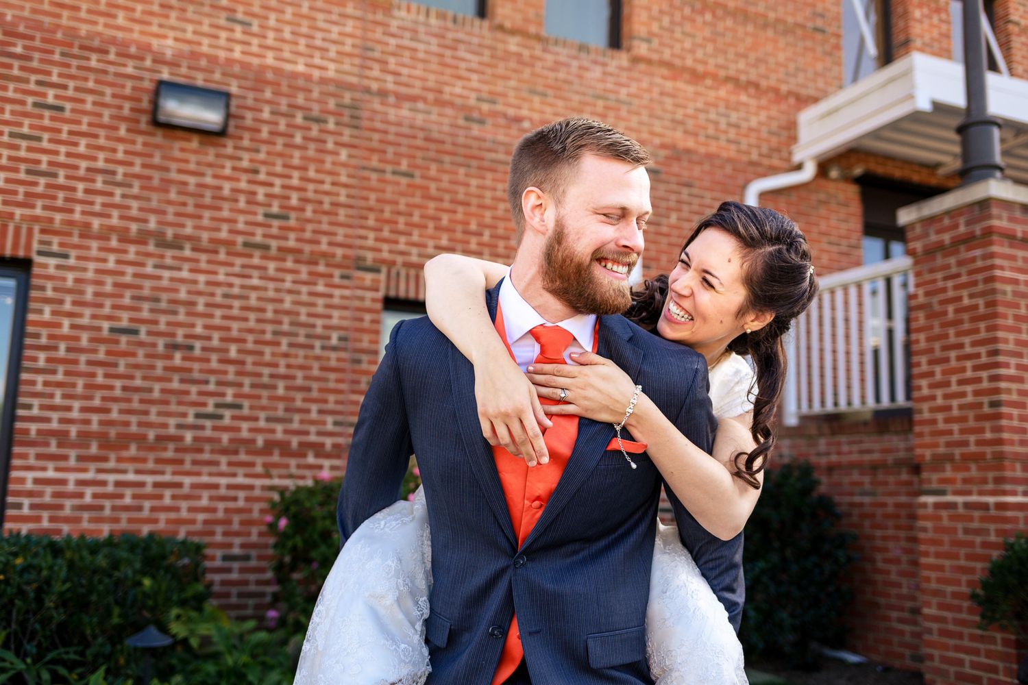 Smiling groom carries bride piggyback outside brick building after wedding ceremony.