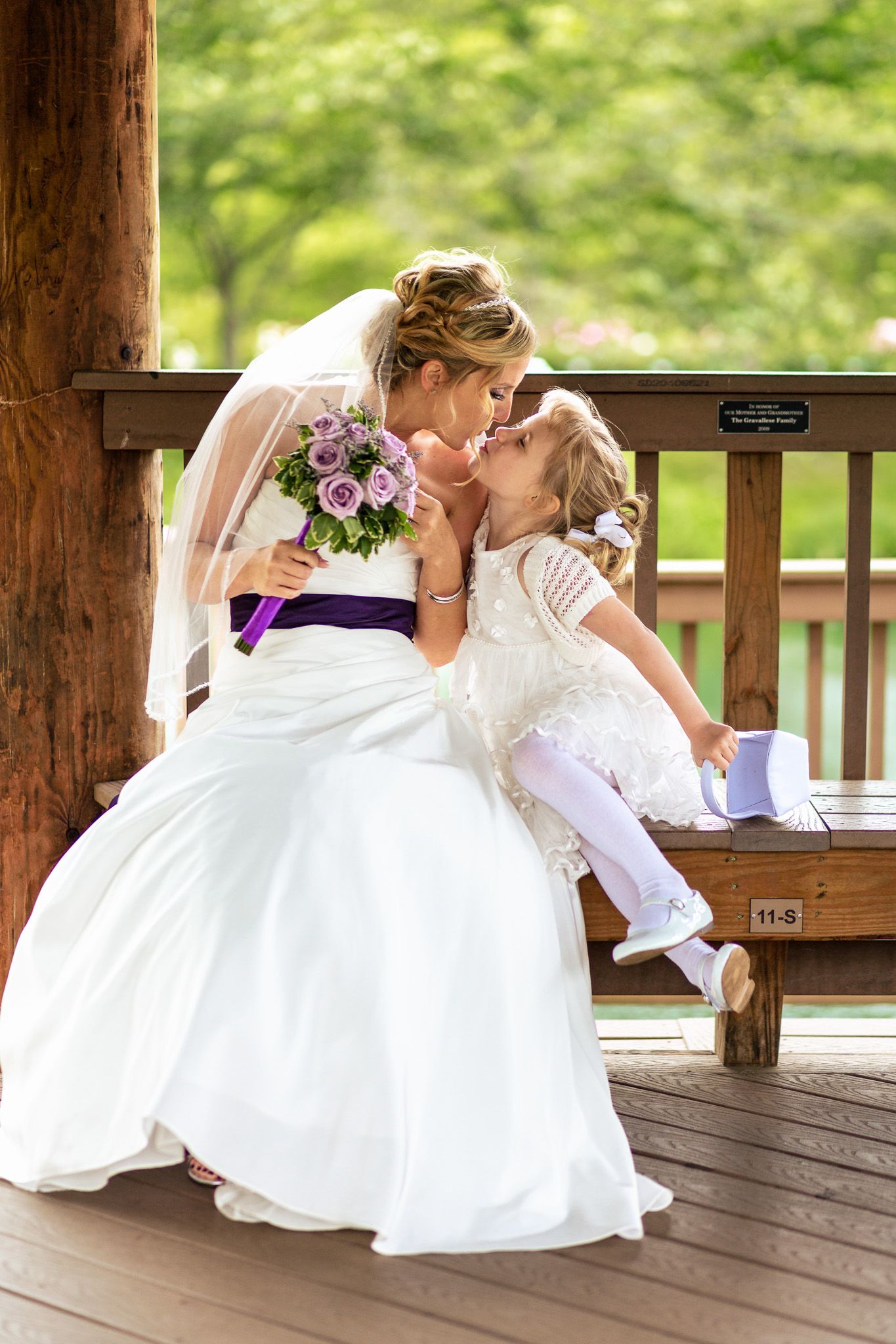 Bride in white gown with purple sash shares a tender kiss with young flower girl on bench.