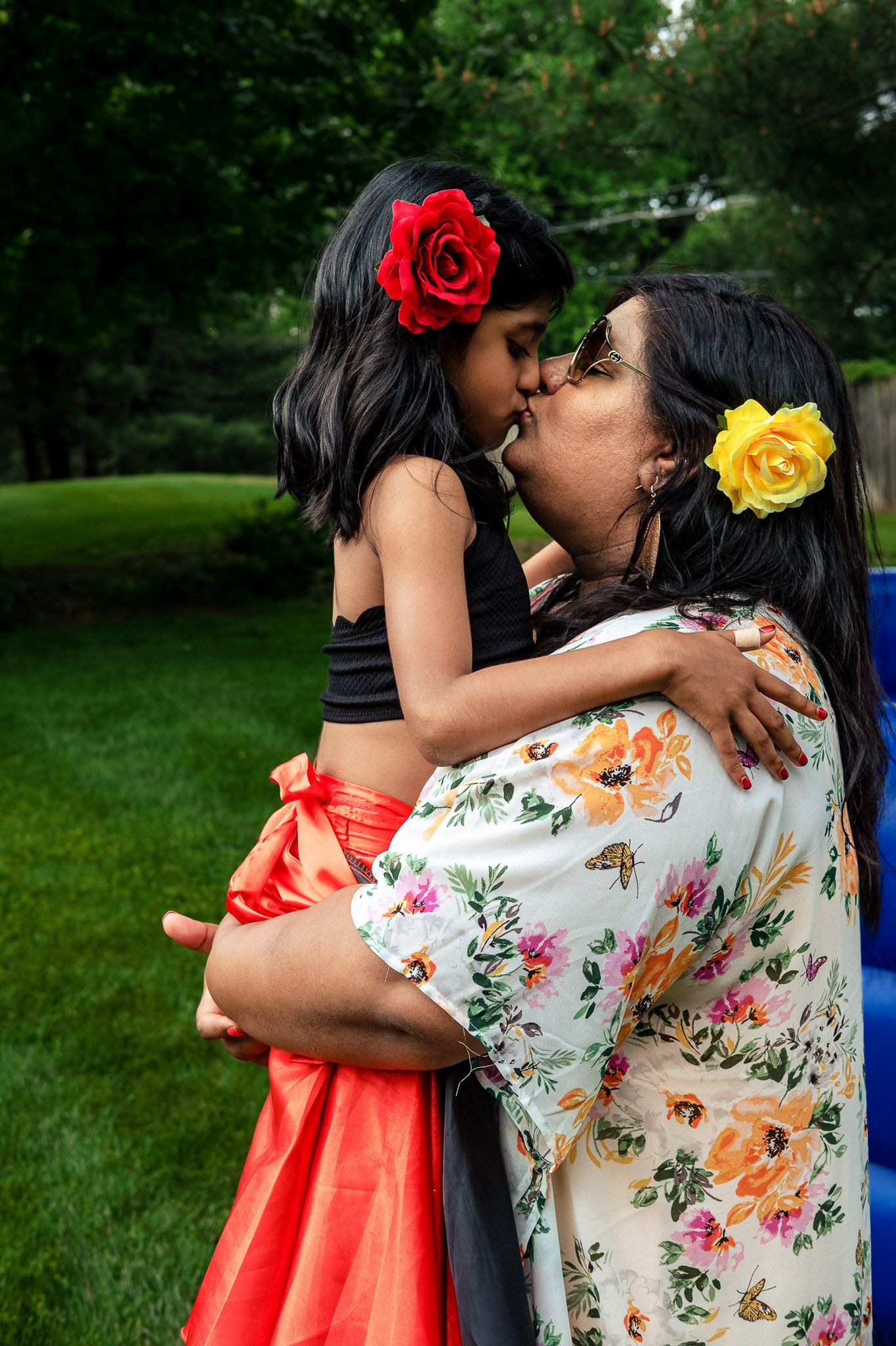 A mother holding and kissing her young daughter, both wearing colorful flowers in their hair outdoors.