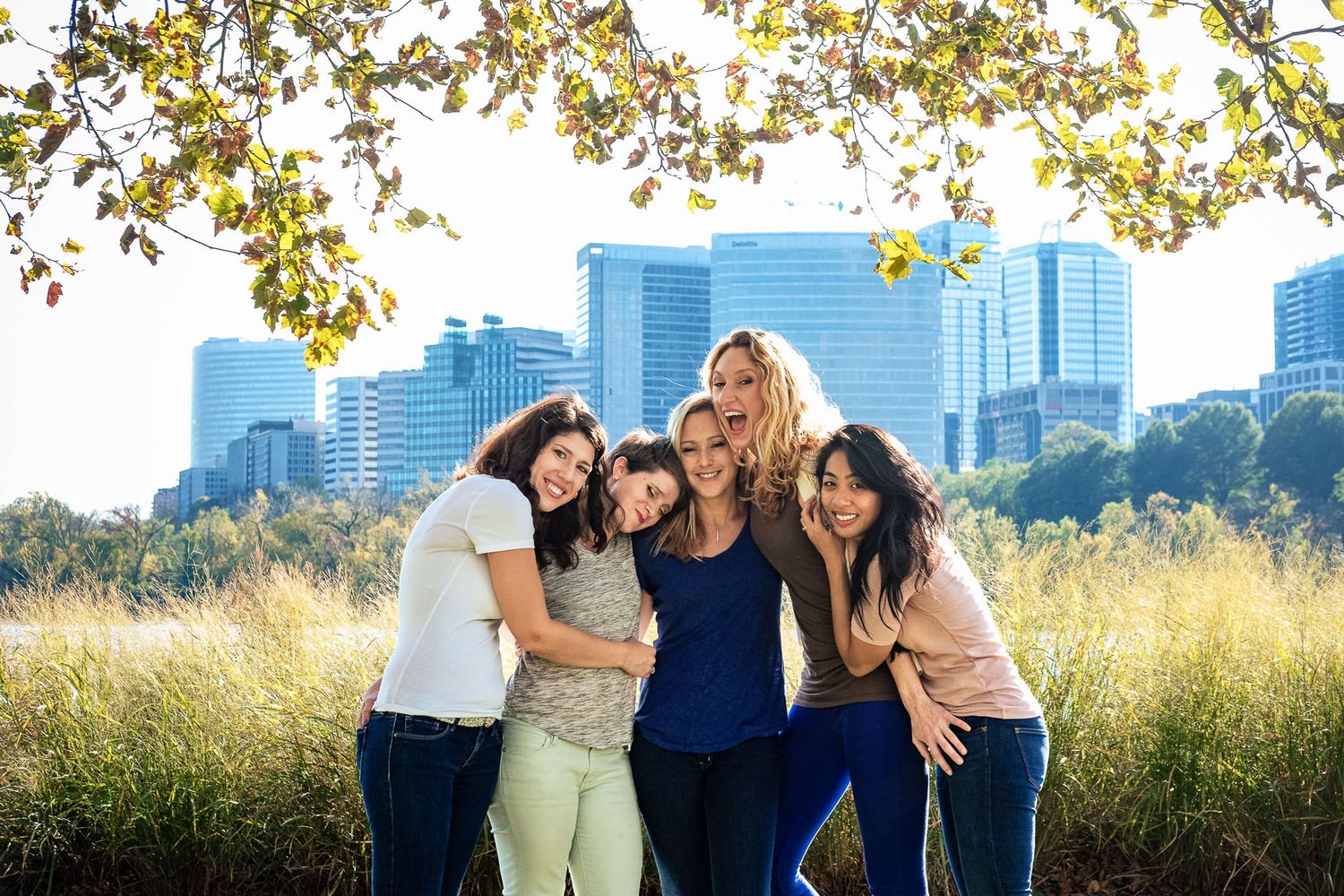 Group of five women hugging and smiling together in urban park with city skyline.