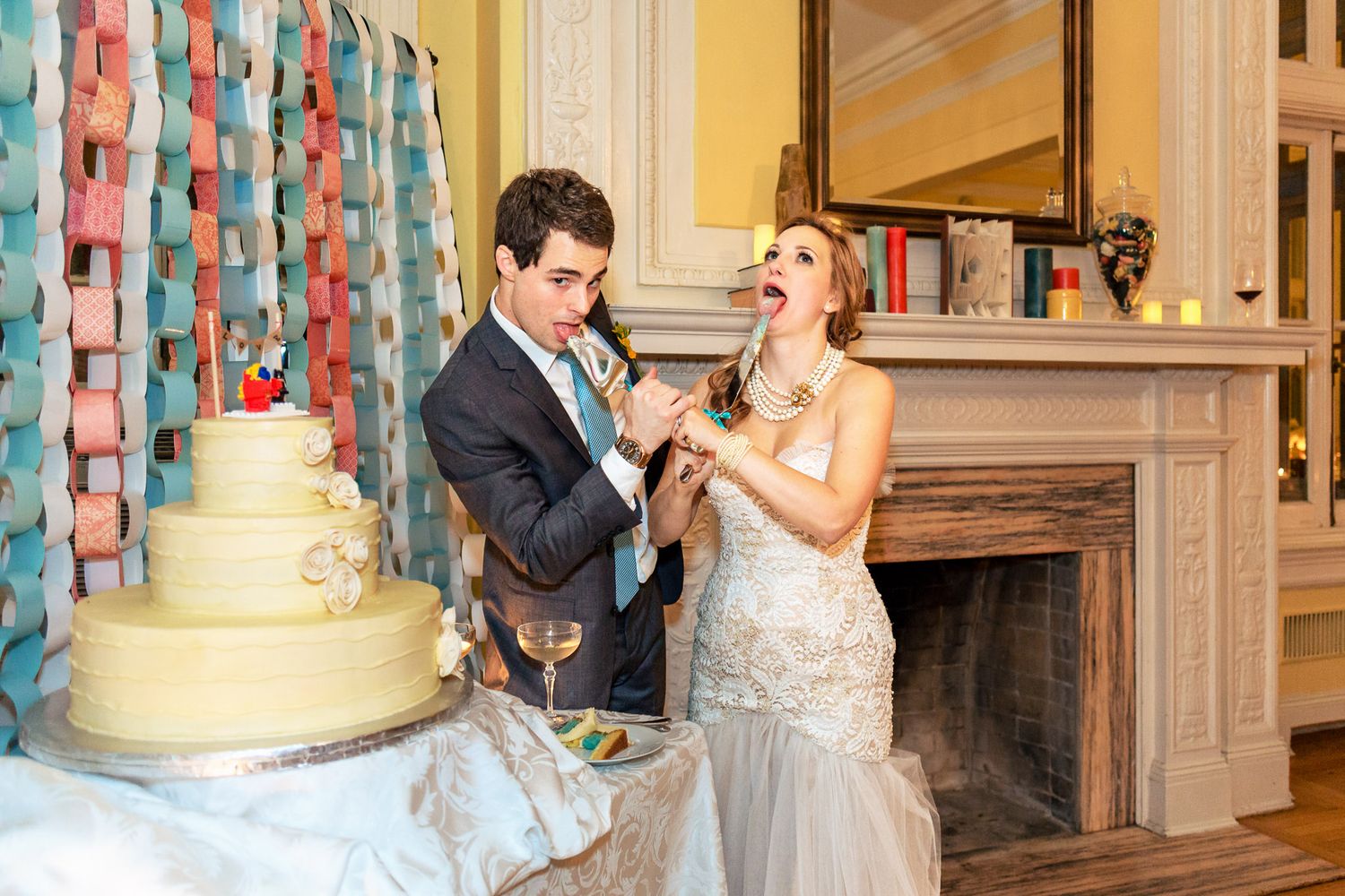 Bride and groom laughing while cutting their white wedding cake near a fireplace at their elegant reception.