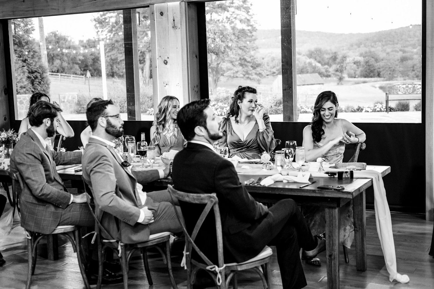 Black and white photo of wedding guests enjoying dinner conversation at rustic barn reception.