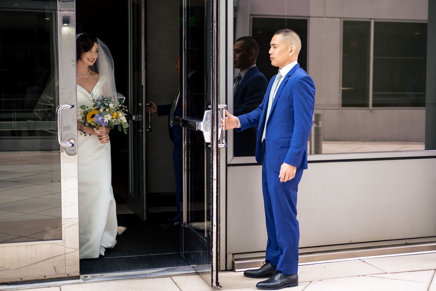 Groom in blue suit holds door open for bride in white wedding dress entering a modern building.