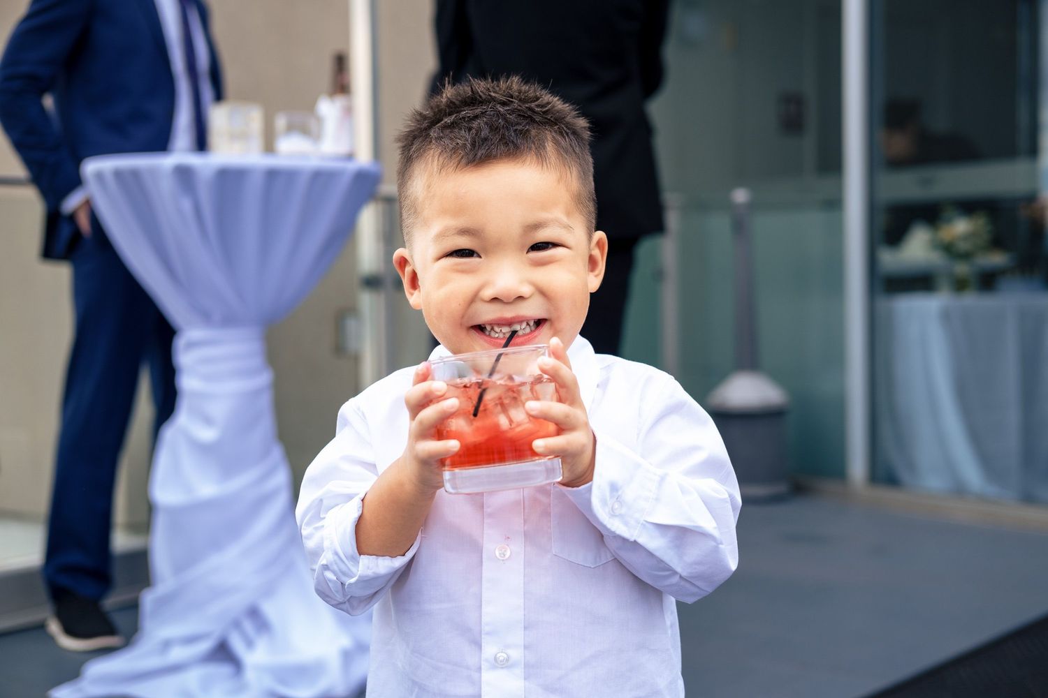 Adorable young boy in white shirt smiles while holding a red drink at an outdoor wedding reception.