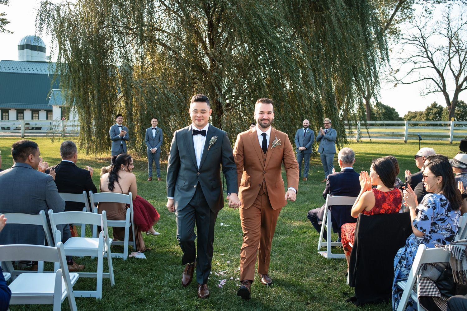 Two grooms walk hand-in-hand down the aisle after their outdoor wedding ceremony past smiling guests.