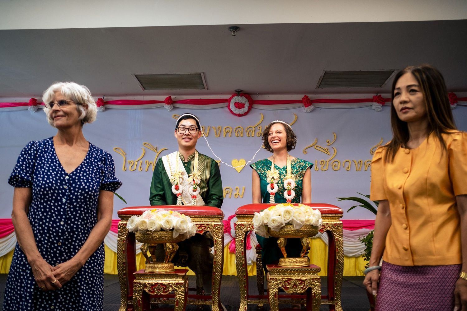 Bride and groom in traditional Thai green wedding outfits seated at ornate golden tables during ceremony.