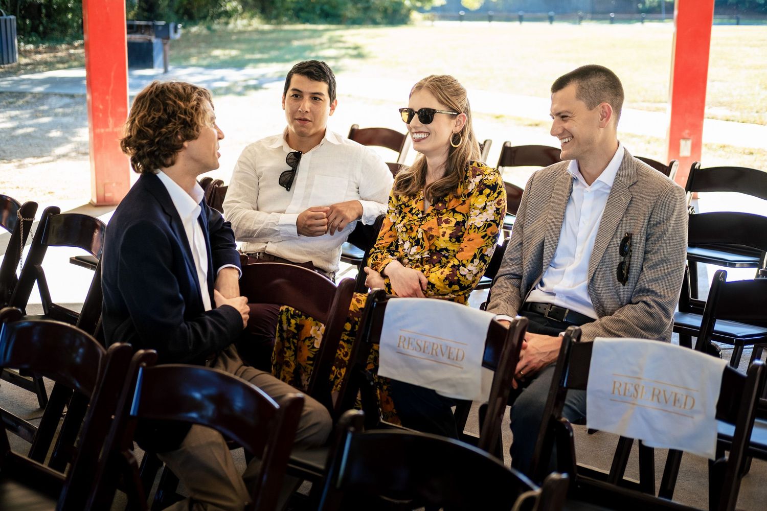 Four wedding guests in formal attire chat and laugh while seated in chairs at an outdoor ceremony venue.