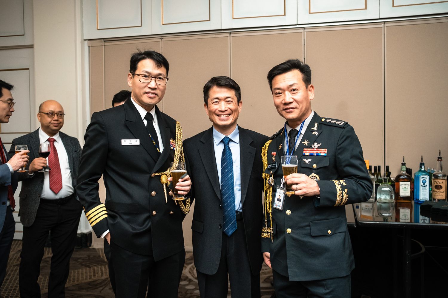 Three men in military and formal attire smile and toast with drinks at an indoor reception event.