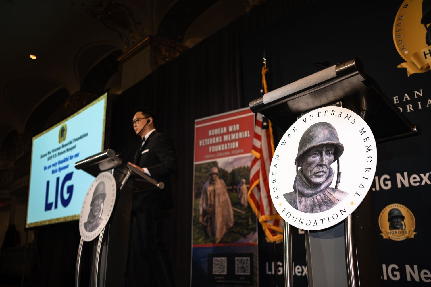 Speaker at podium during Korean War Veterans Memorial Foundation event with LIG banner and memorial logo displays.