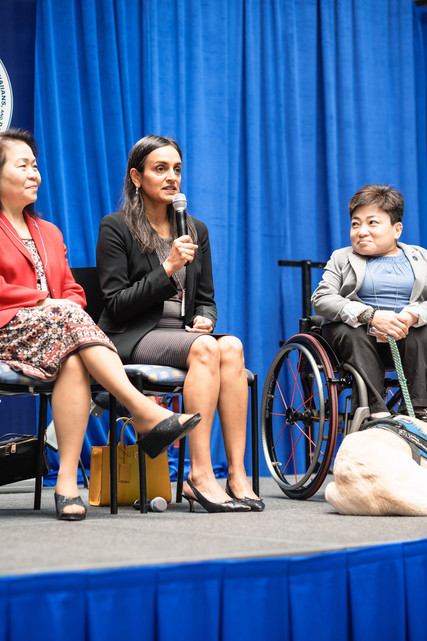 Three women on stage at a panel event, one speaking into a microphone, another in a wheelchair with a service dog nearby.
