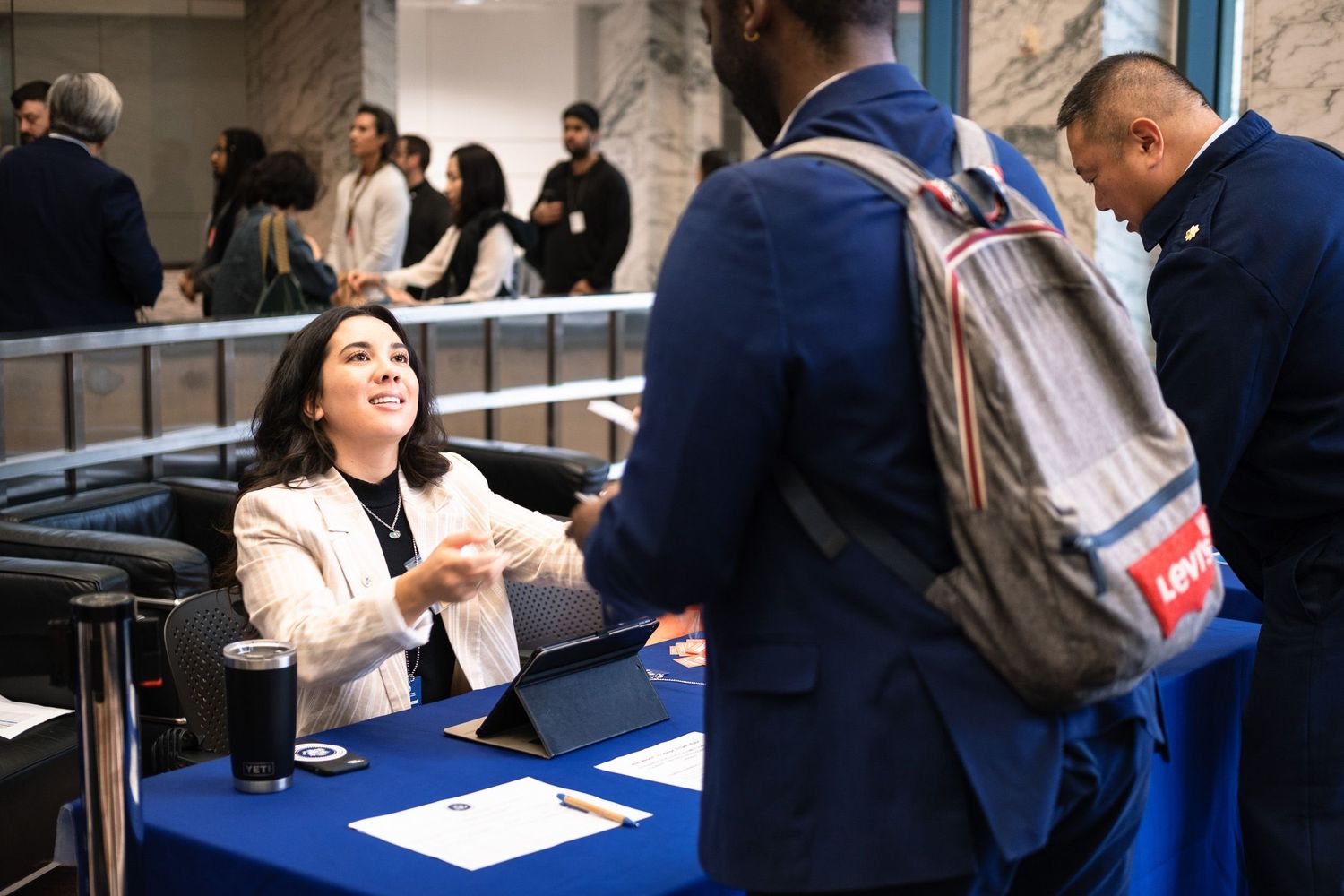 A smiling woman at a blue table shaking hands with a visitor at a professional event.