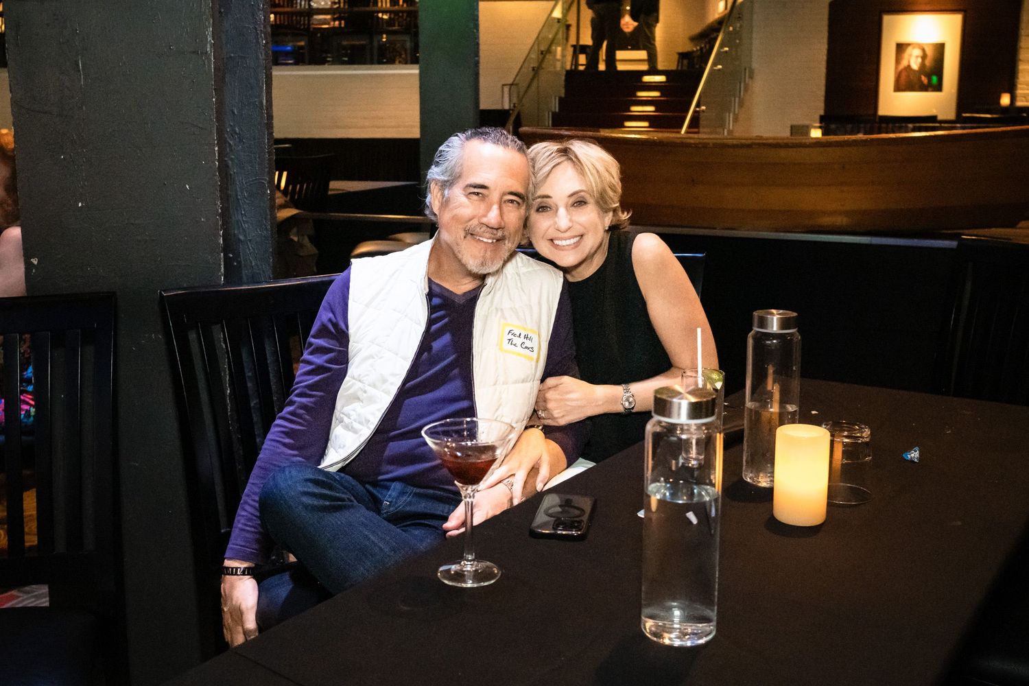 Two smiling guests pose together at a dimly lit restaurant table with candles and drinks during a social event.