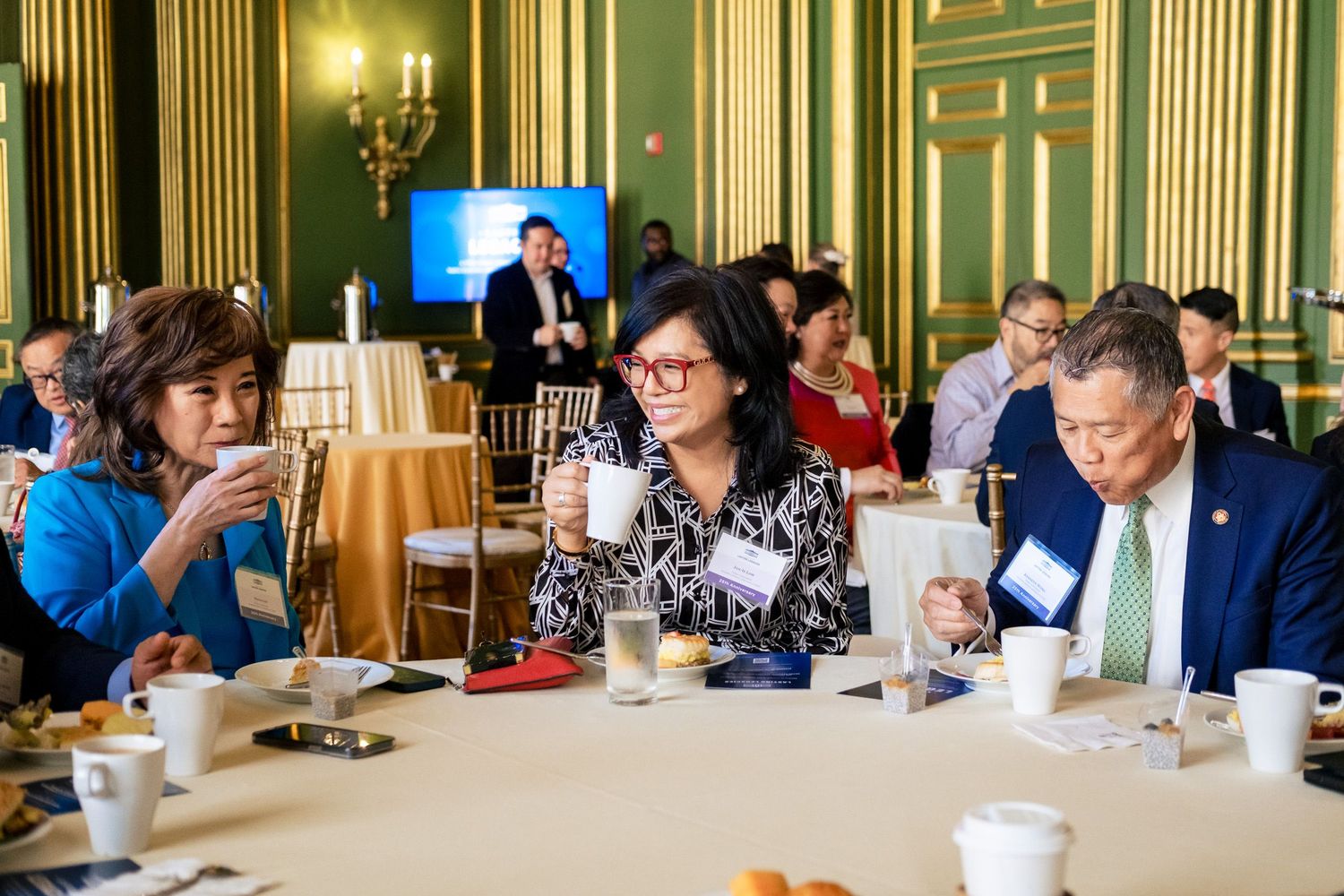 Three attendees converse over coffee at a formal event in a grand green and gold ballroom.