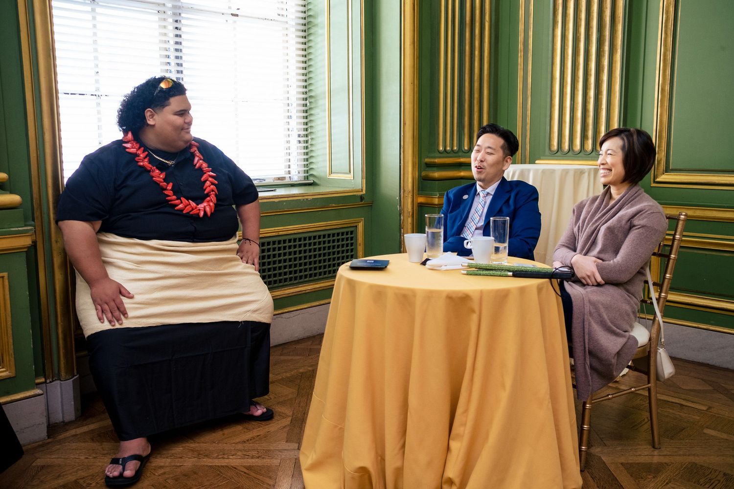 Three people in elegant green room, one wearing floral lei, two seated at gold-draped table in formal meeting setting.