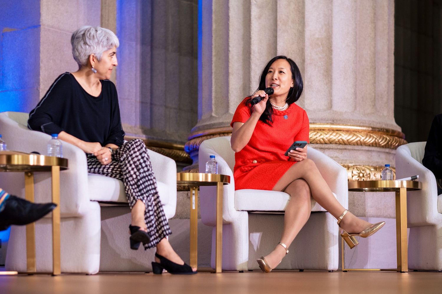 Two women in elegant attire engage in a panel discussion on a modern white stage with gold accents and marble columns.