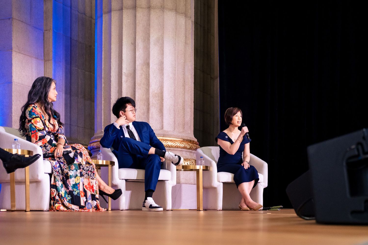 Three panelists seated on stage at a formal conference event with large columns in the background.