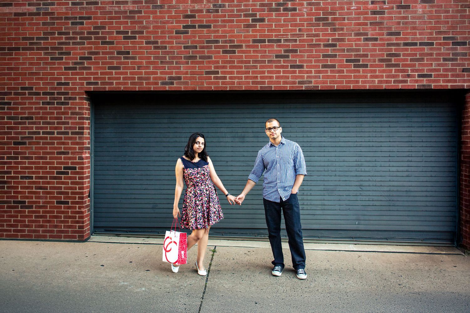 Couple holding hands in front of a brick wall and metal garage door on a sidewalk.
