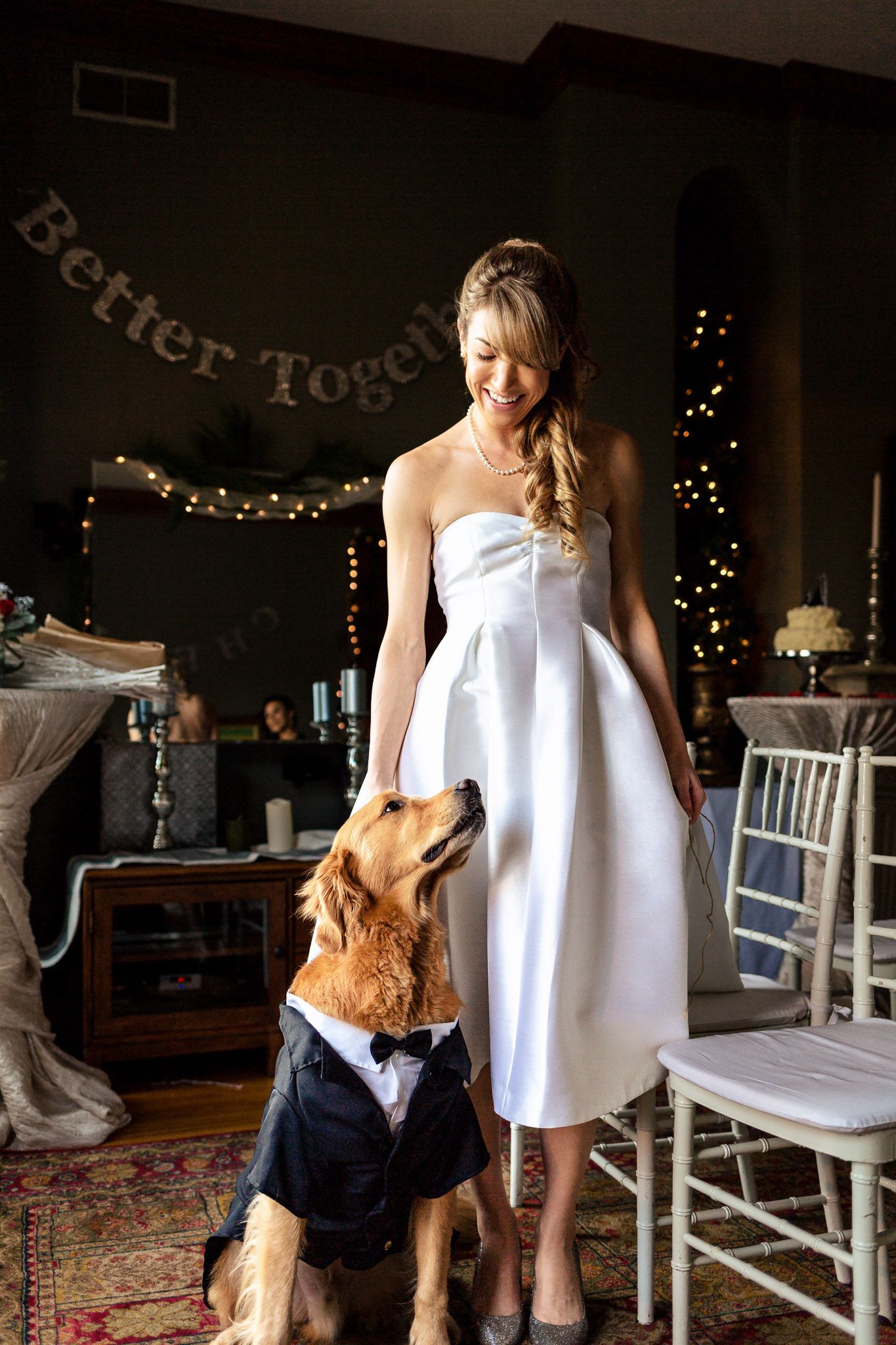 Smiling bride in white dress stands beside a golden retriever dressed in a tuxedo.
