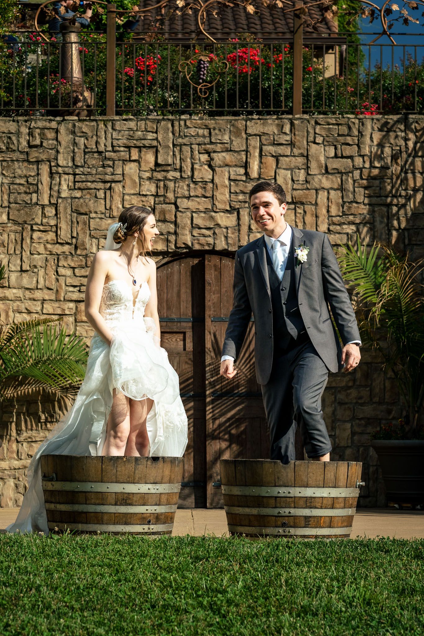 Bride and groom smile at each other while standing in wooden wine barrels outside a rustic stone venue.