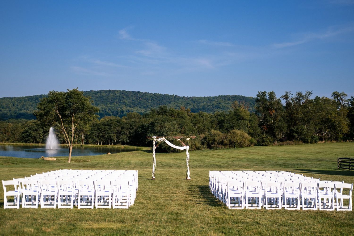 Outdoor wedding ceremony setup with white chairs, floral arch, pond fountain, and mountain views.