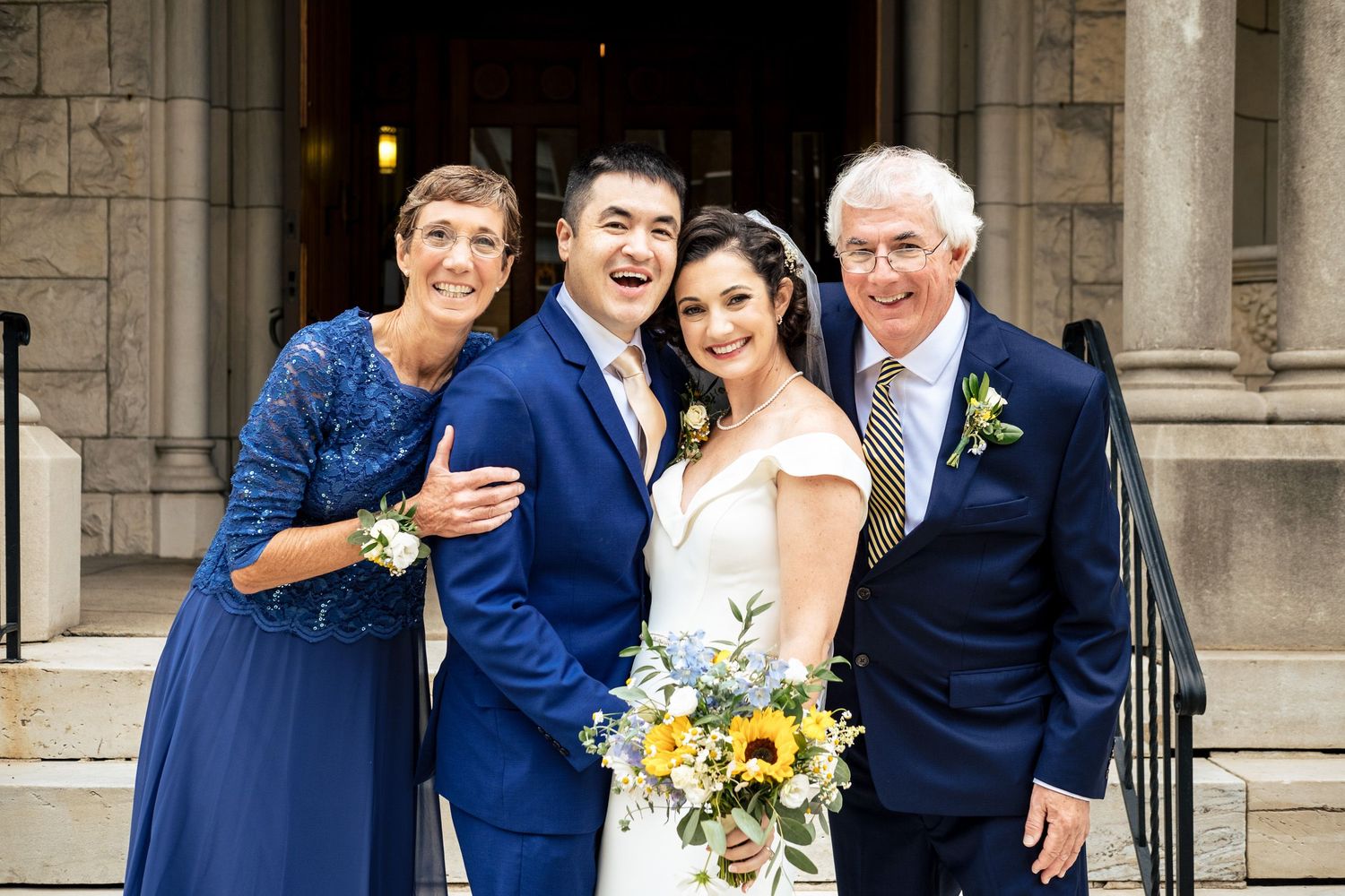 Bride and groom in blue suit posing joyfully with parents in blue lace and sunflower bouquet on church steps.