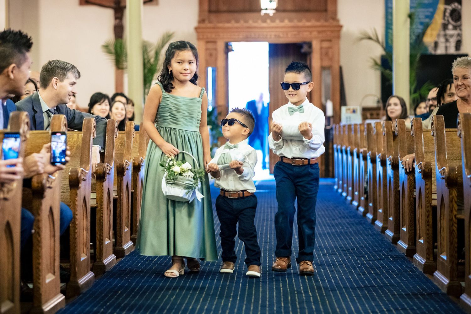 Three children walk down a church aisle, wearing sunglasses, as flower girl and ring bearers at a wedding ceremony.