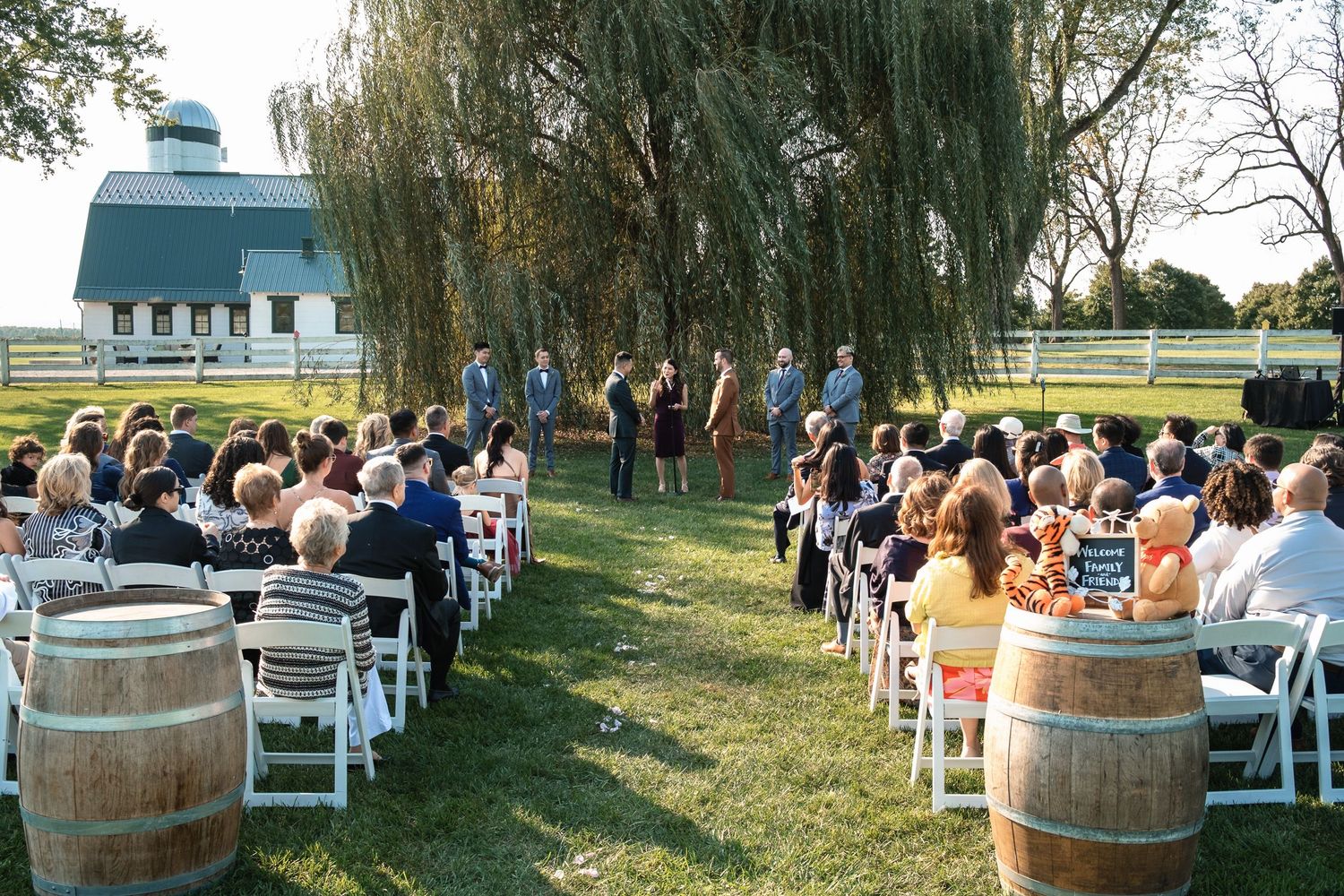 Guests seated at an outdoor farm wedding ceremony beside a blue barn under a weeping willow tree.