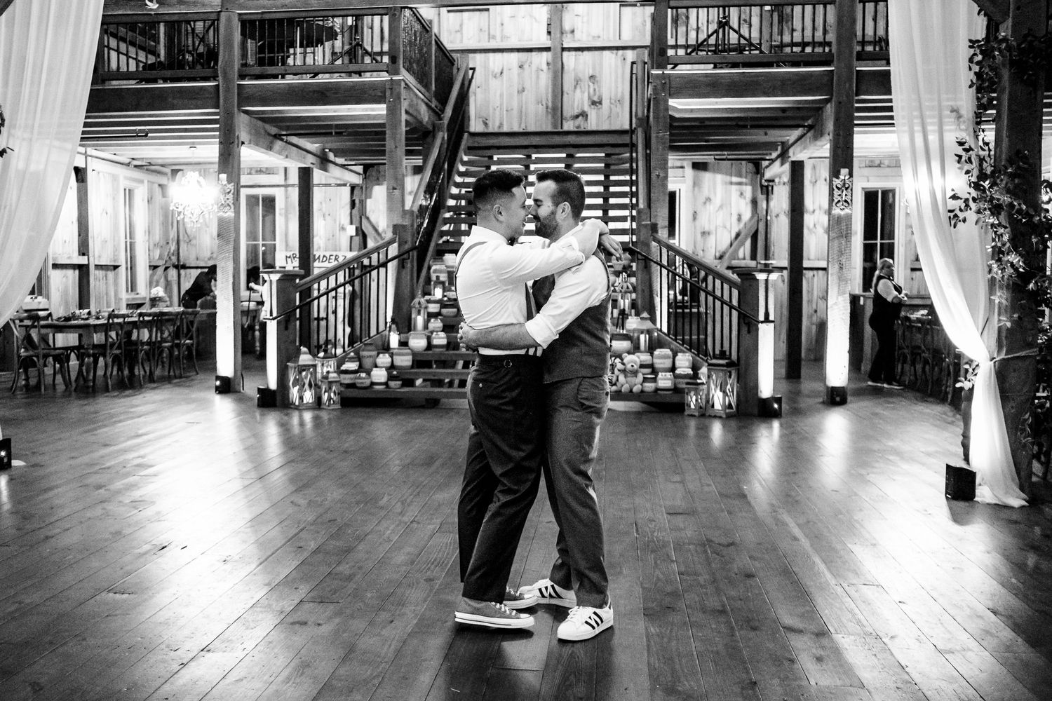 Two grooms share an intimate first dance on a hardwood floor in a rustic barn wedding.