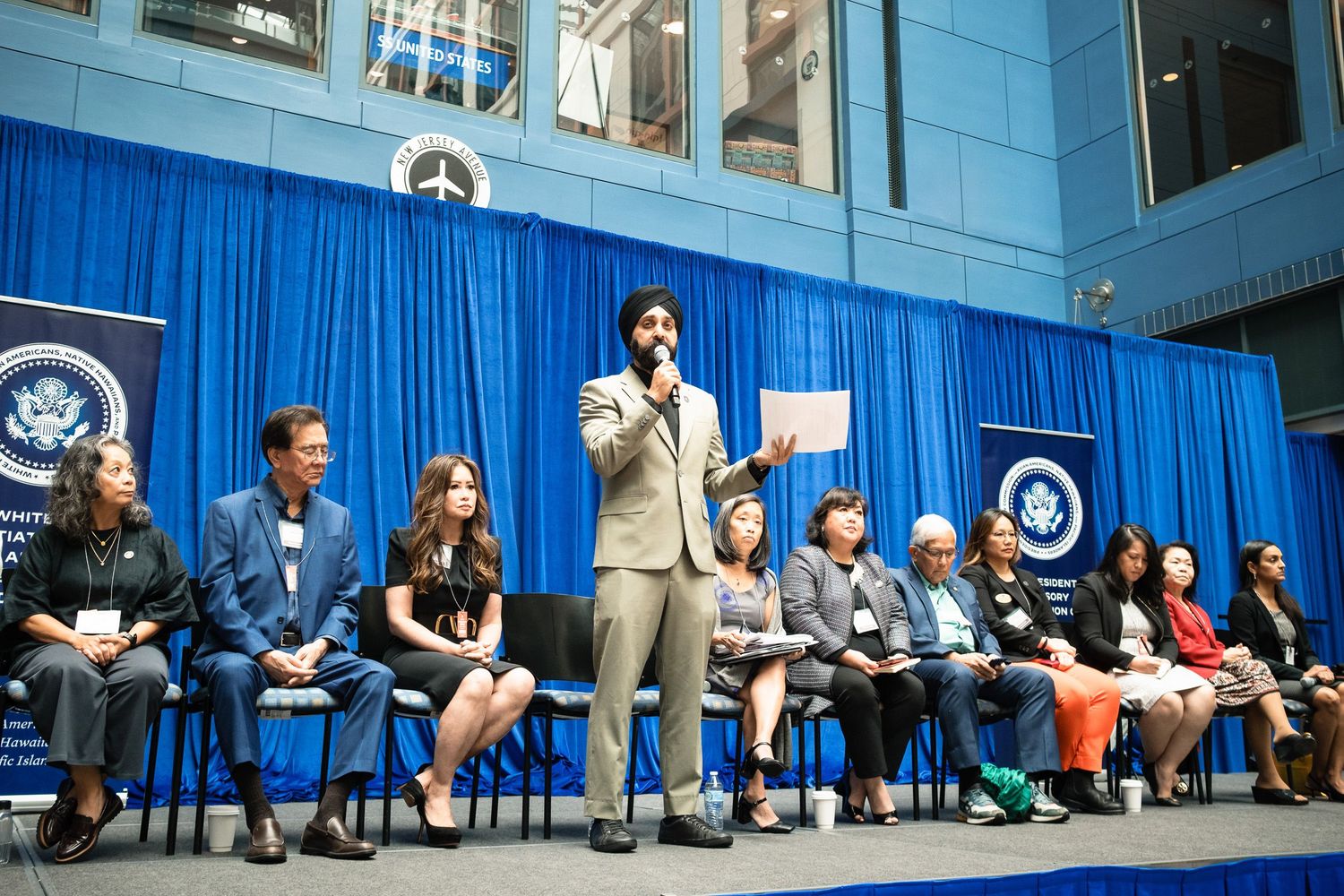 Speaker addresses seated panel at formal event with blue curtain and official seal backdrop indoors.