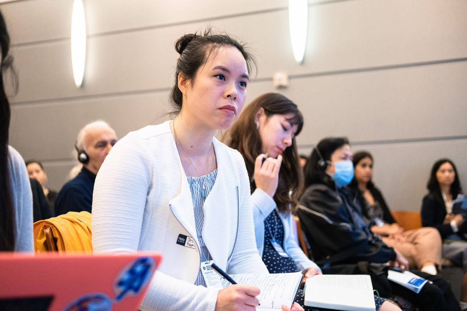 Attentive woman in white blazer taking notes at a professional conference with attendees in background.