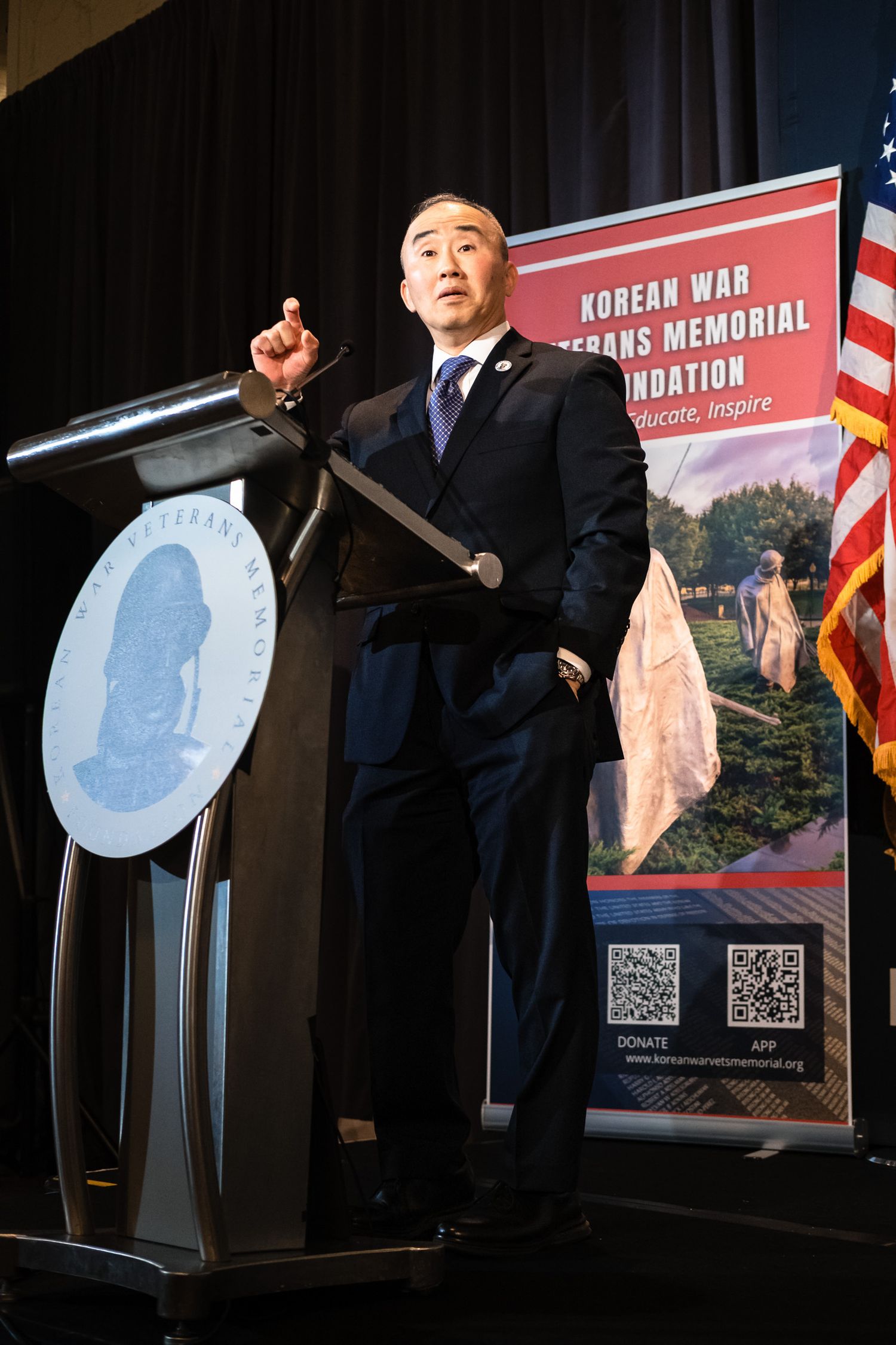 Man in suit speaks at podium for Korean War Veterans Memorial Foundation event with banner displayed behind him.