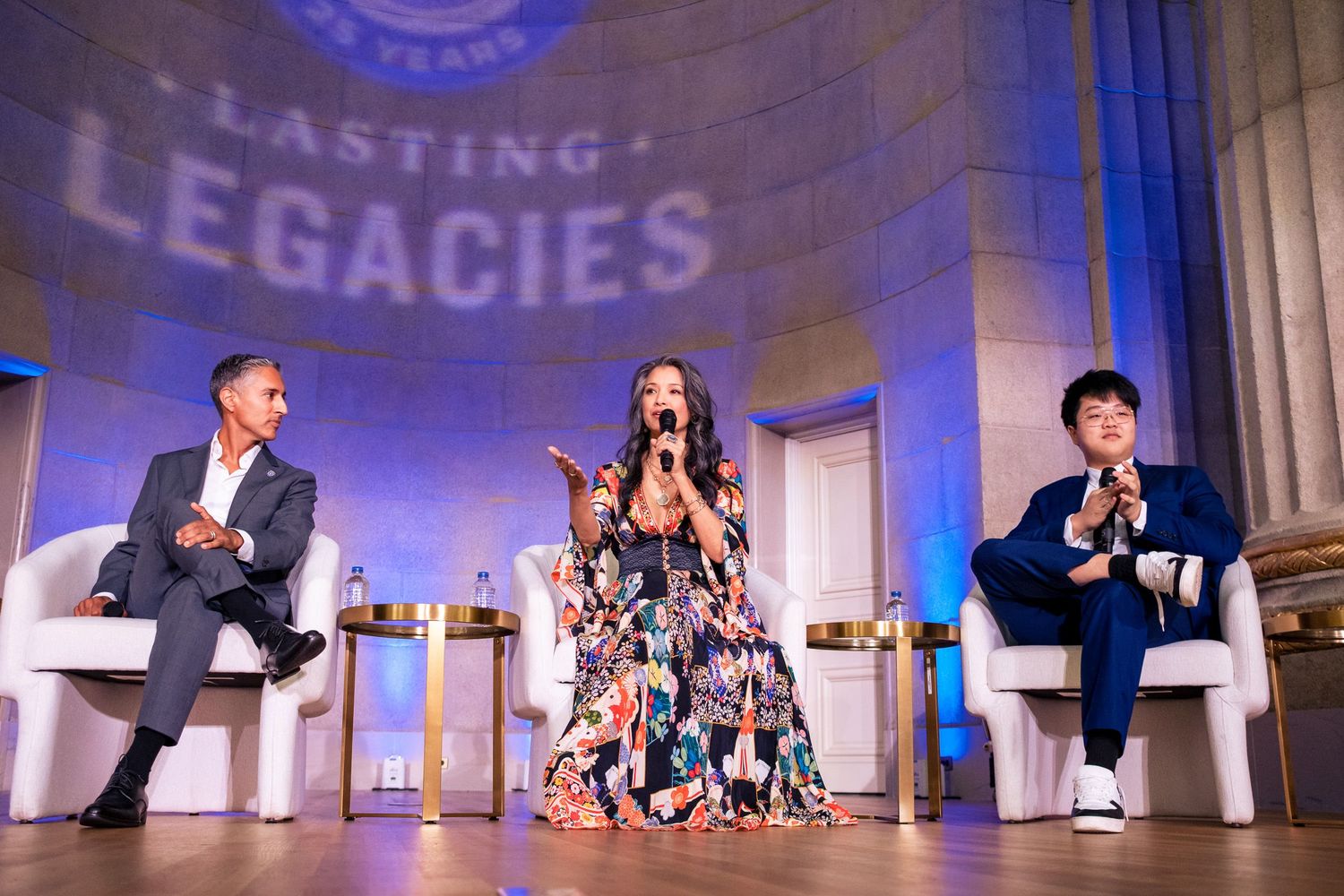 Three speakers engage in panel discussion at Lasting Legacies anniversary event on a blue-lit stage.