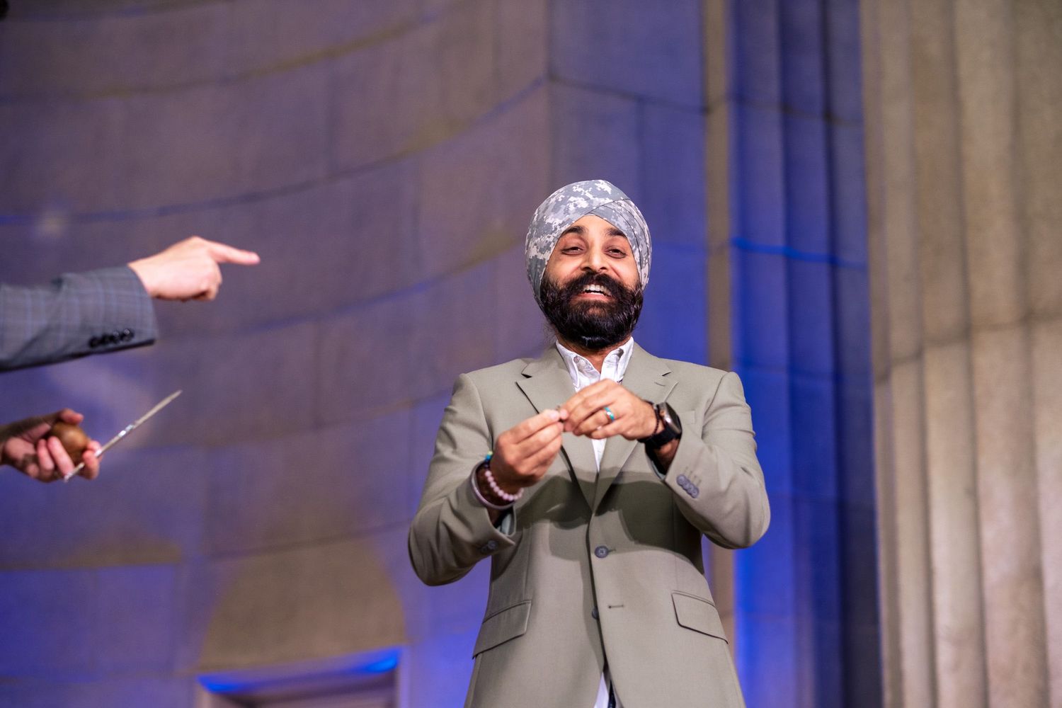 Bearded man in gray suit and turban smiles while holding object on stage with dramatic column backdrop.