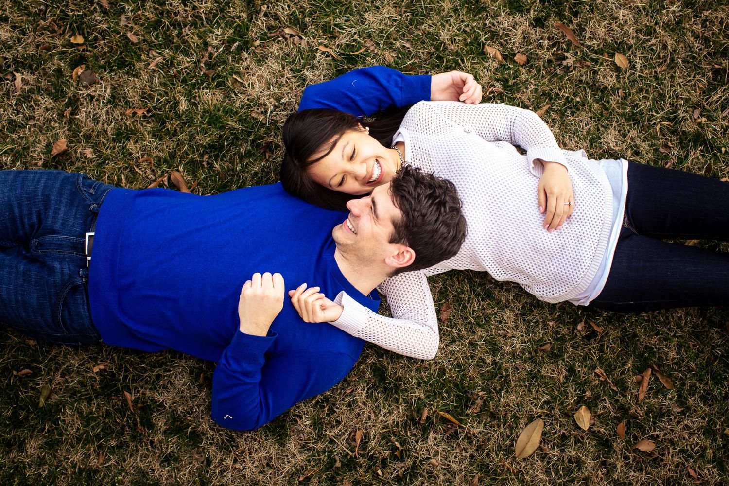 Couple lying on grass in a park, facing each other and holding hands from above.