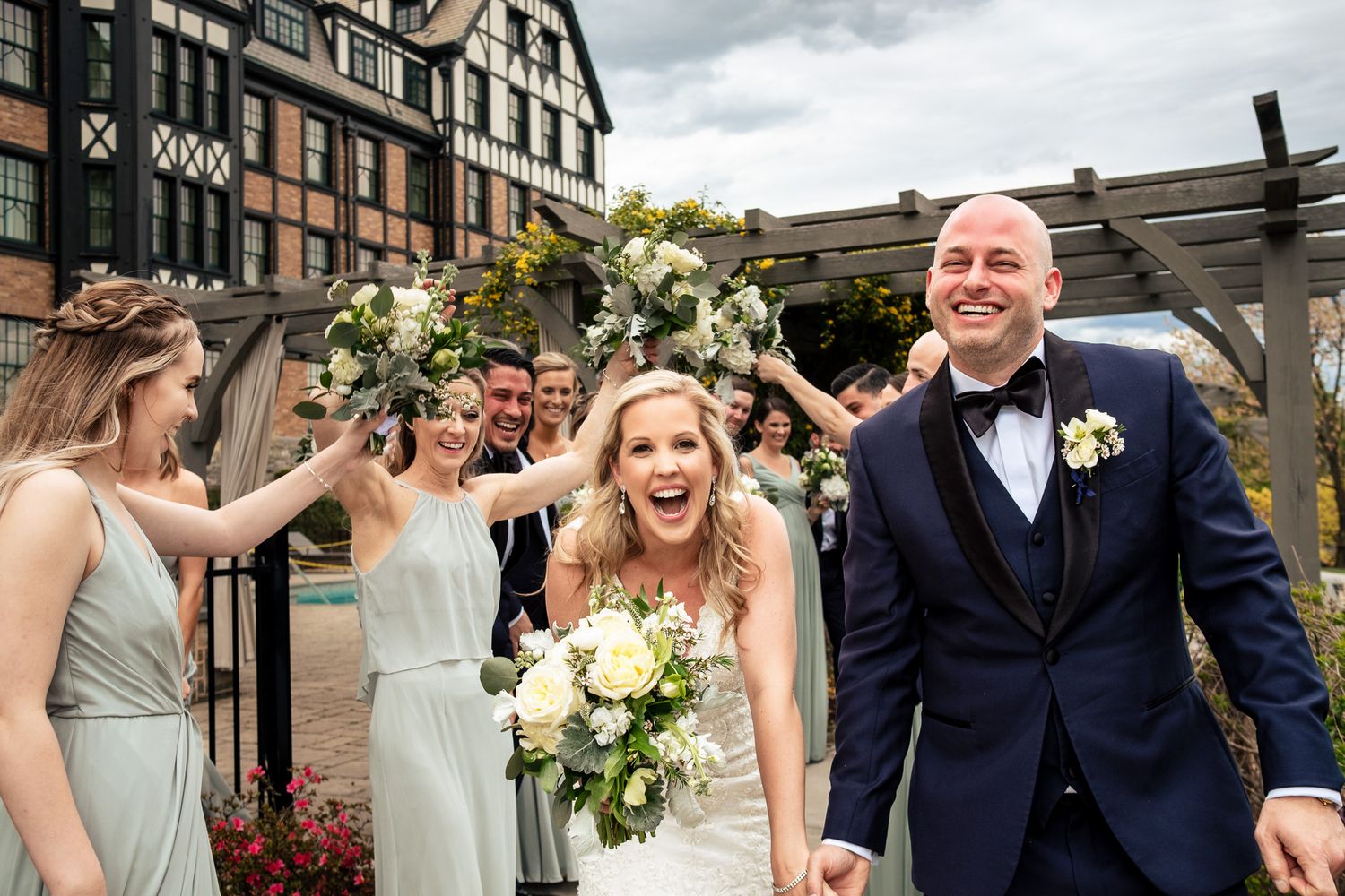 Ecstatic bride and groom walk through floral arch as bridesmaids celebrate at outdoor wedding.