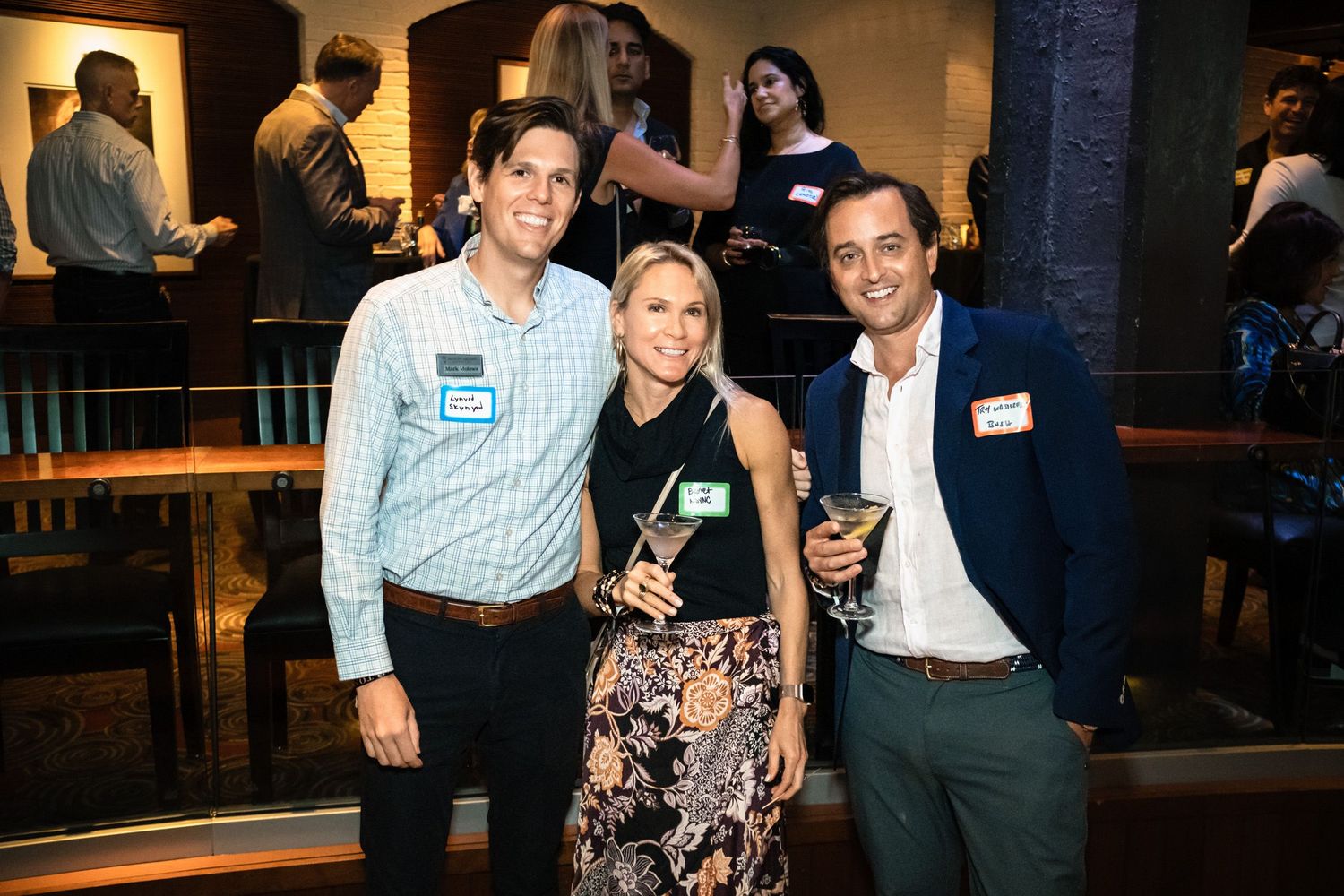 Three adults smile and pose with drinks at a lively indoor networking event or party.