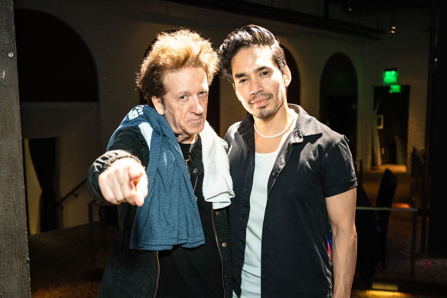 Two men posing together indoors, one pointing at camera, wearing casual outfits at an event.