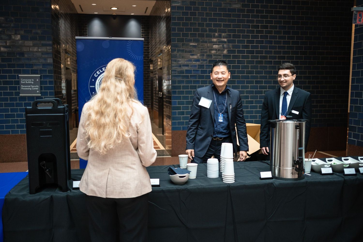 Two men smile at a woman at a registration table during a professional networking event in a modern venue.